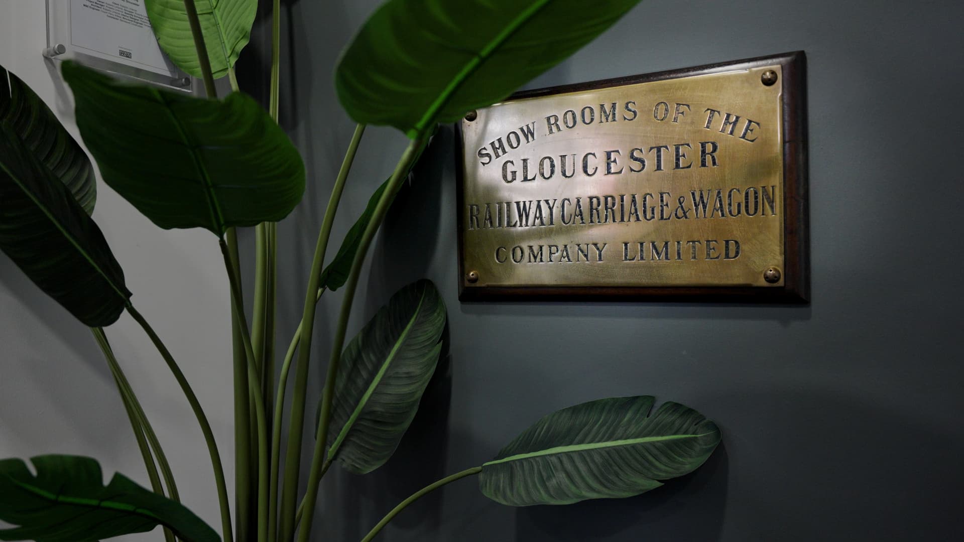 Historic brass plaque reading “Show rooms of the Gloucester Railway Carriage & Wagon Company Limited”, mounted on a dark wall beside a plant